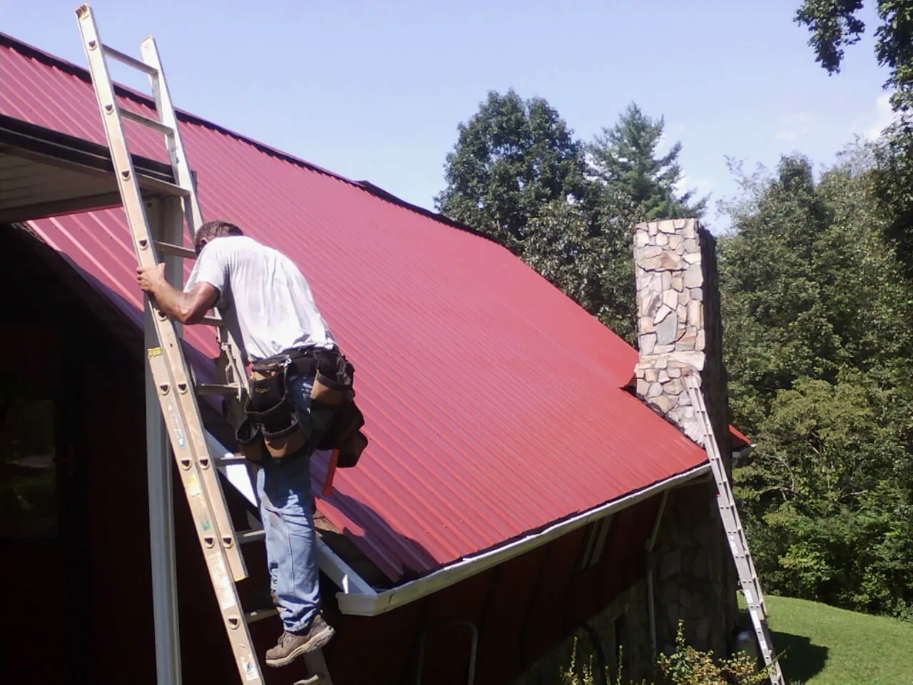 Roofer on a ladder beside a red metal roof and stone chimney — Soffit & Fascia Repair by Roofing Brothers in Chiloquin
